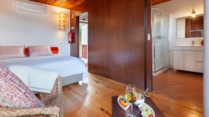 A bedroom with a bed, a wicker chair, a dining table with fruit and wine, and a kitchen in the background. - CASA DO RANCHO (Amares)