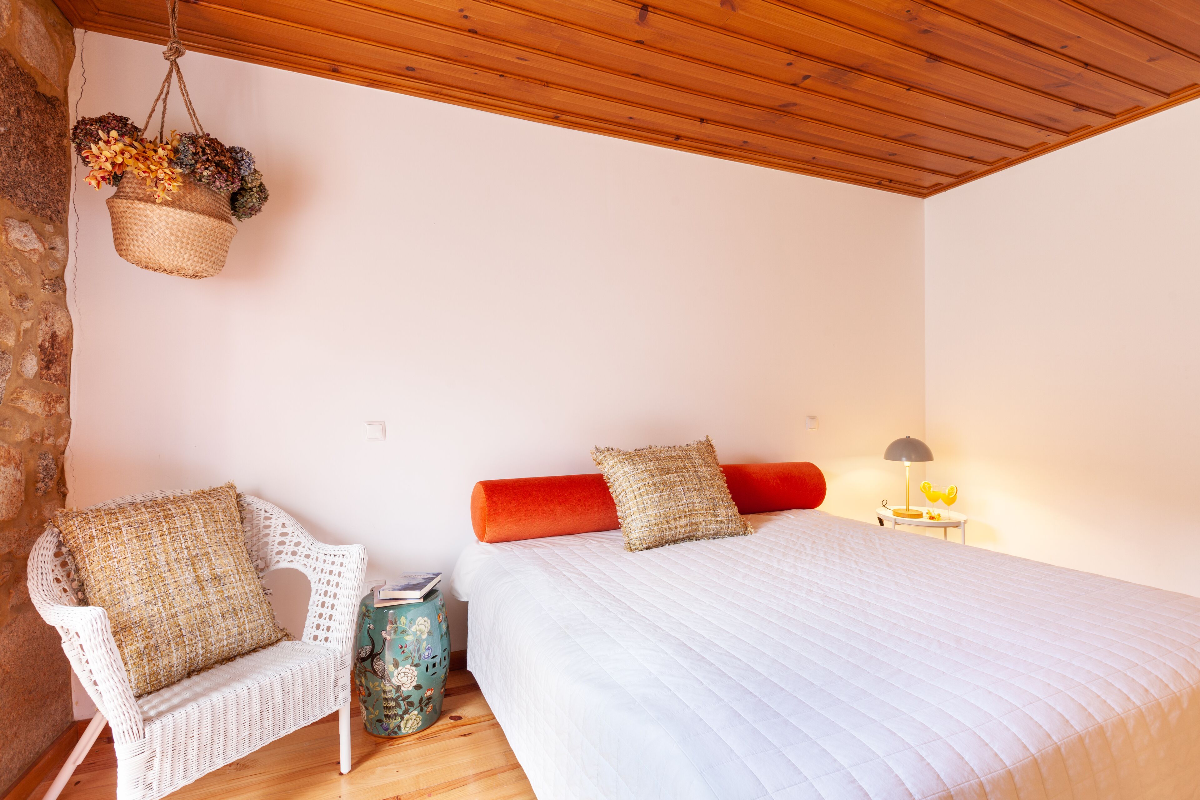 A bedroom with a white bed, two pillows, a wicker chair, a small table with a lamp, and a hanging basket with dried flowers.