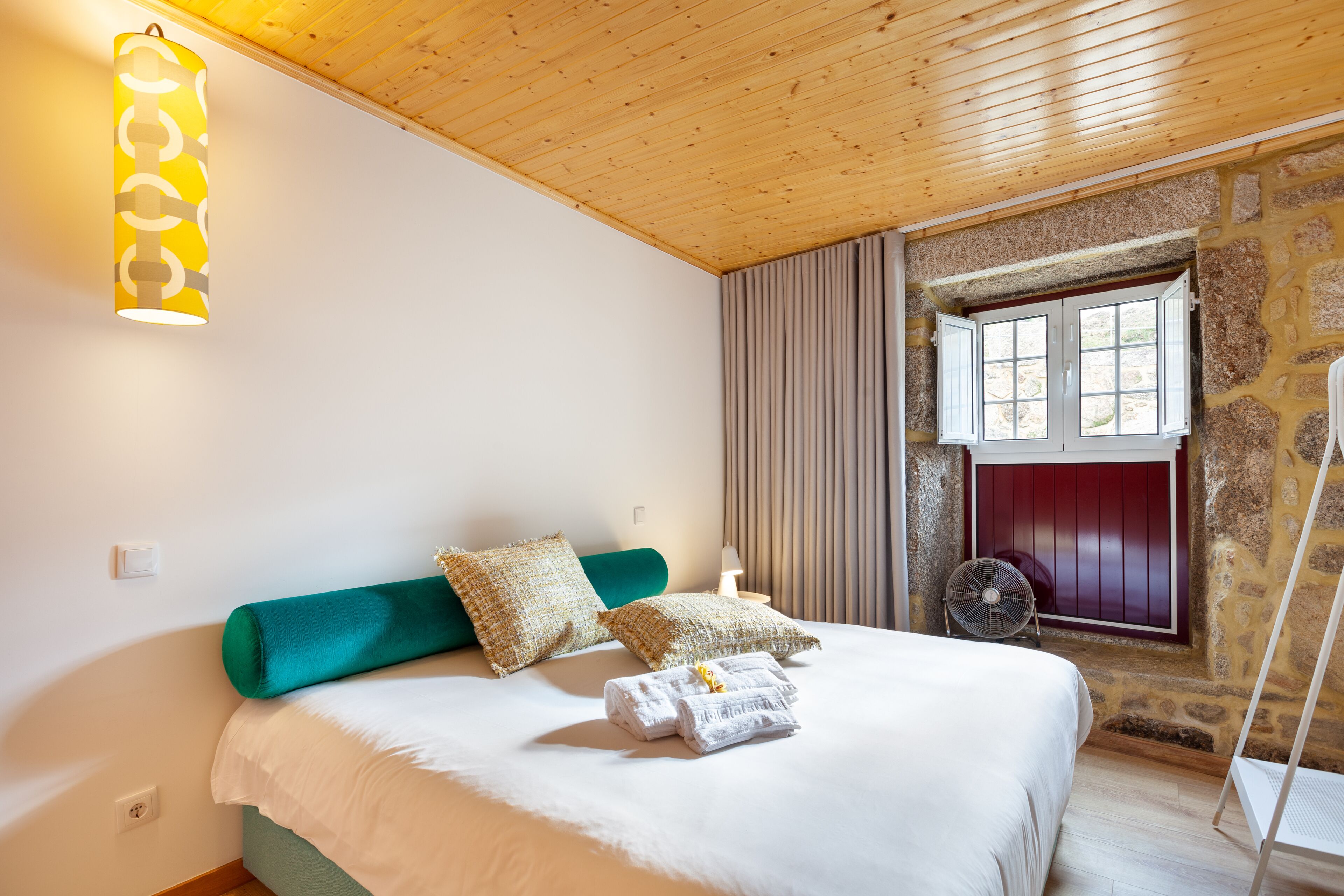 A bedroom with a bed, pillows, a bedside table, a lamp, a fan, and a window with curtains.