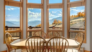 A dining area with a round table and wooden chairs, large windows with a view of mountains and autumn foliage.