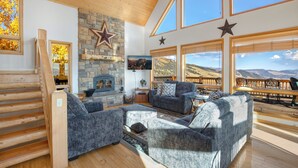 A spacious living room with a stone fireplace, comfortable seating, large windows with mountain views, and a wooden ceiling.
