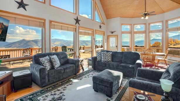A spacious living room with large windows offering mountain views, a high wooden ceiling, and a cozy seating area with a fireplace.