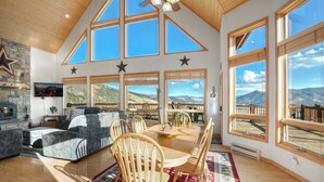 A spacious living room with a high ceiling, large windows offering mountain views, a fireplace, and a dining area with wooden furniture.