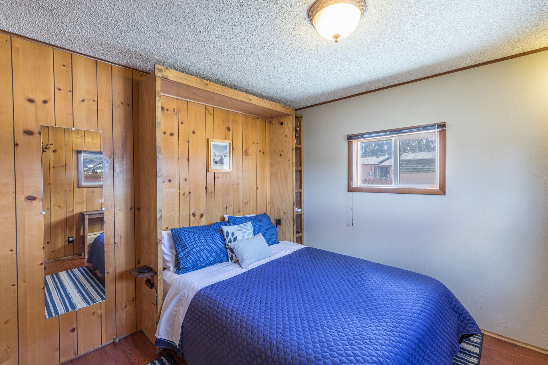 A bedroom with a wooden panel wall, a bed with blue bedding, a framed picture on the wall, and a window with a view.