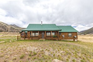 A rustic log cabin with a green roof, a porch, and a mountain backdrop. - Creede Cabin - Dogs Welcome - Amazing Views - Large Deck (Creede)