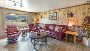 Una sala de estar acogedora con un sofá de cuero rojo, dos sillones, una mesa de centro de madera y vistas a un paisaje montañoso a través de un ventanal.