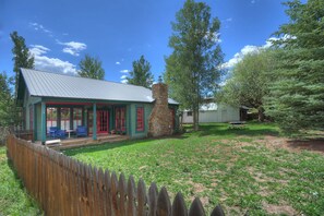A rustic cabin with a stone chimney, a porch with blue chairs, and a wooden fence.