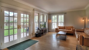 A spacious living room with a leather sofa, a coffee table, and a TV on a stand. Large windows provide a view of the outdoors. - Historic Home | Pet Friendly | Walkable to Downtown (Creede)