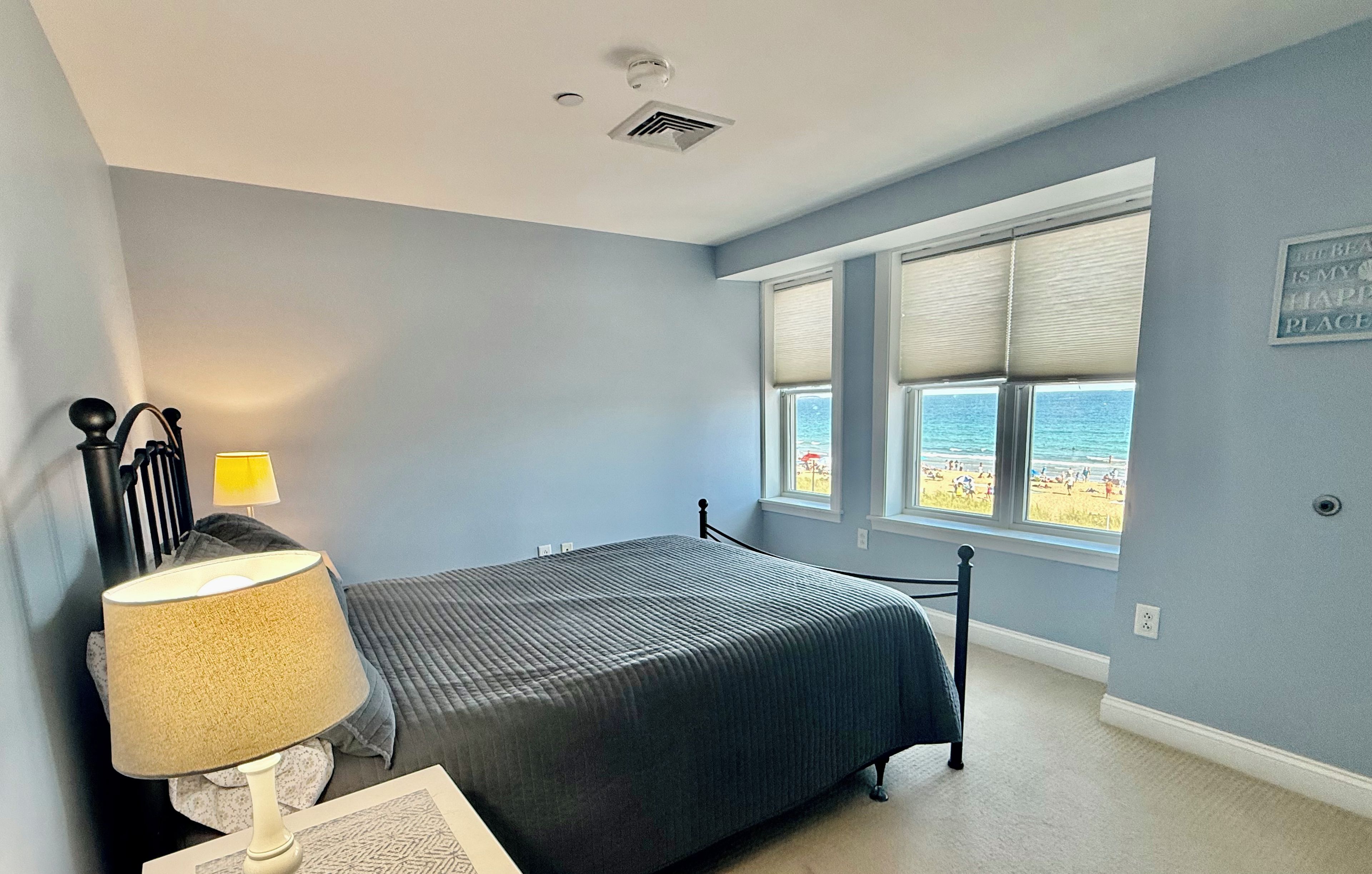 A bedroom with a bed, a nightstand, a lamp, a window with a view of the beach, and a ceiling vent.