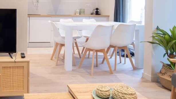 A modern living room with a dining area, a TV, and a kitchen in the background.
