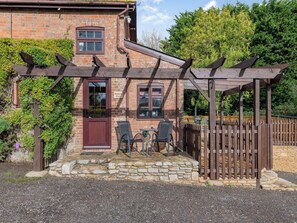 A brick building with a wooden pergola, stone steps, and two chairs outside.