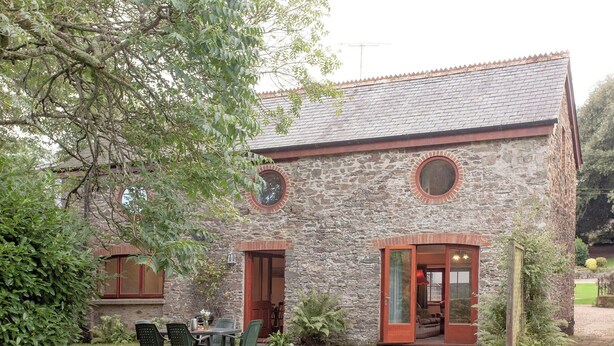 A stone building with a tiled roof, a round window, and a door leading to a covered porch. - Coachman's lodge - UK35952 (Kingsbridge)