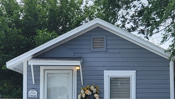 A small, single-story building with a gray exterior and white trim. It has a front door with the number 213, a small porch with a white railing, and a wreath above the door. - THE COZY COTTAGE, A Great place to hang your hat. (Post)