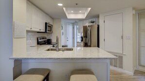A modern kitchen with white cabinets, a stainless steel refrigerator, a microwave, and a large island with bar stools. - 206 Barrington Arms- OCEANFRONT and BEAUTIFUL (Hilton Head Island)