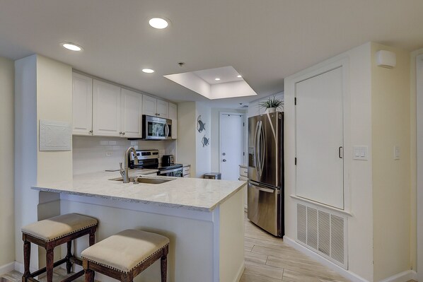 Una cocina moderna con armarios blancos, electrodomésticos de acero inoxidable, una isla central con taburetes e iluminación empotrada en el techo.
