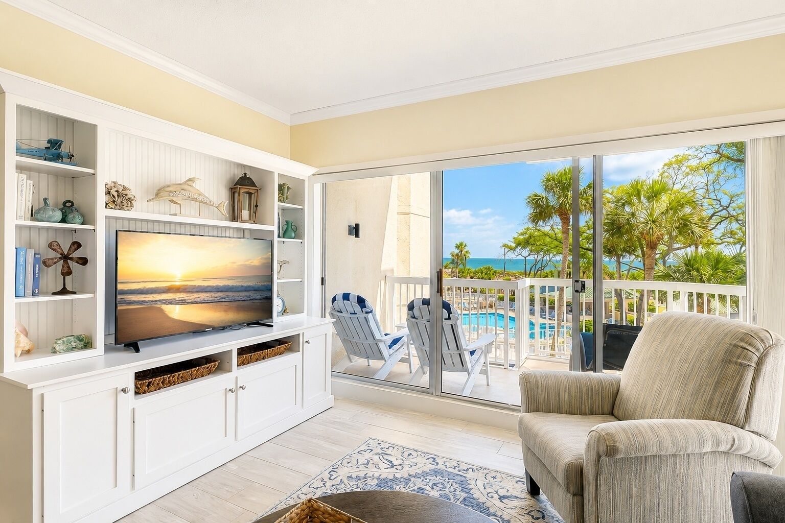 A living room with a TV, white shelves, a pool, and a balcony with a view of the ocean.