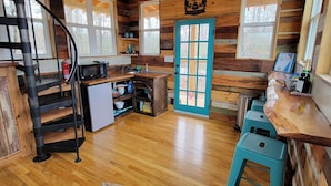 Interior de una cabaña con paredes de madera, una kitchenette, una puerta azul y una escalera de caracol.