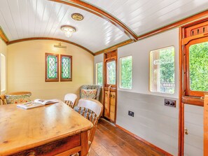 Un coin repas avec une table et des chaises en bois, une horloge au mur et des fenêtres donnant sur un paysage verdoyant.