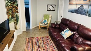 A living room with a leather sofa, a colorful rug, a TV stand, and wall art including a sunset scene and a palm tree. - Sealy Suites B Historic East End - Walk to Beach, UTMB & Strand (Galveston)