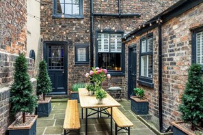 A courtyard with a wooden table and benches, surrounded by brick walls and potted plants.