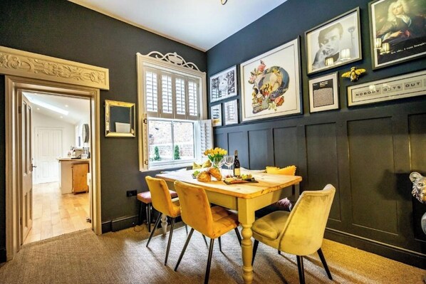 A dining area with a wooden table and yellow chairs, a window with white shutters, and framed pictures on the wall.