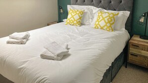 A neatly made bed with white linens and two yellow patterned pillows, a grey upholstered headboard, and a bedside table with a lamp.