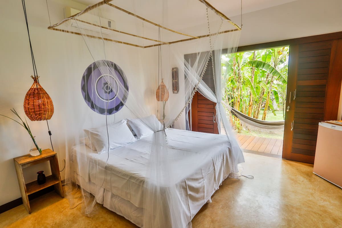 A bedroom with a bed under a mosquito net, a bedside table with a lamp, and a view of a garden through an open door.
