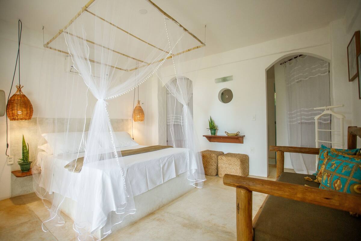 A bedroom with a bed under a mosquito net, a wooden chair, a small table with a plant, and a mirror.