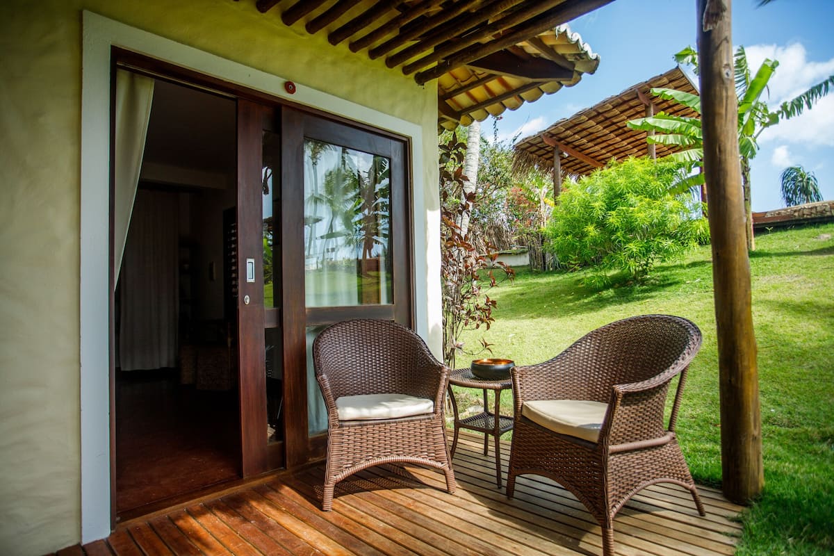 A wooden deck with wicker chairs and a table, a sliding glass door leading to an interior room, and a lush garden with a thatched roof structure.