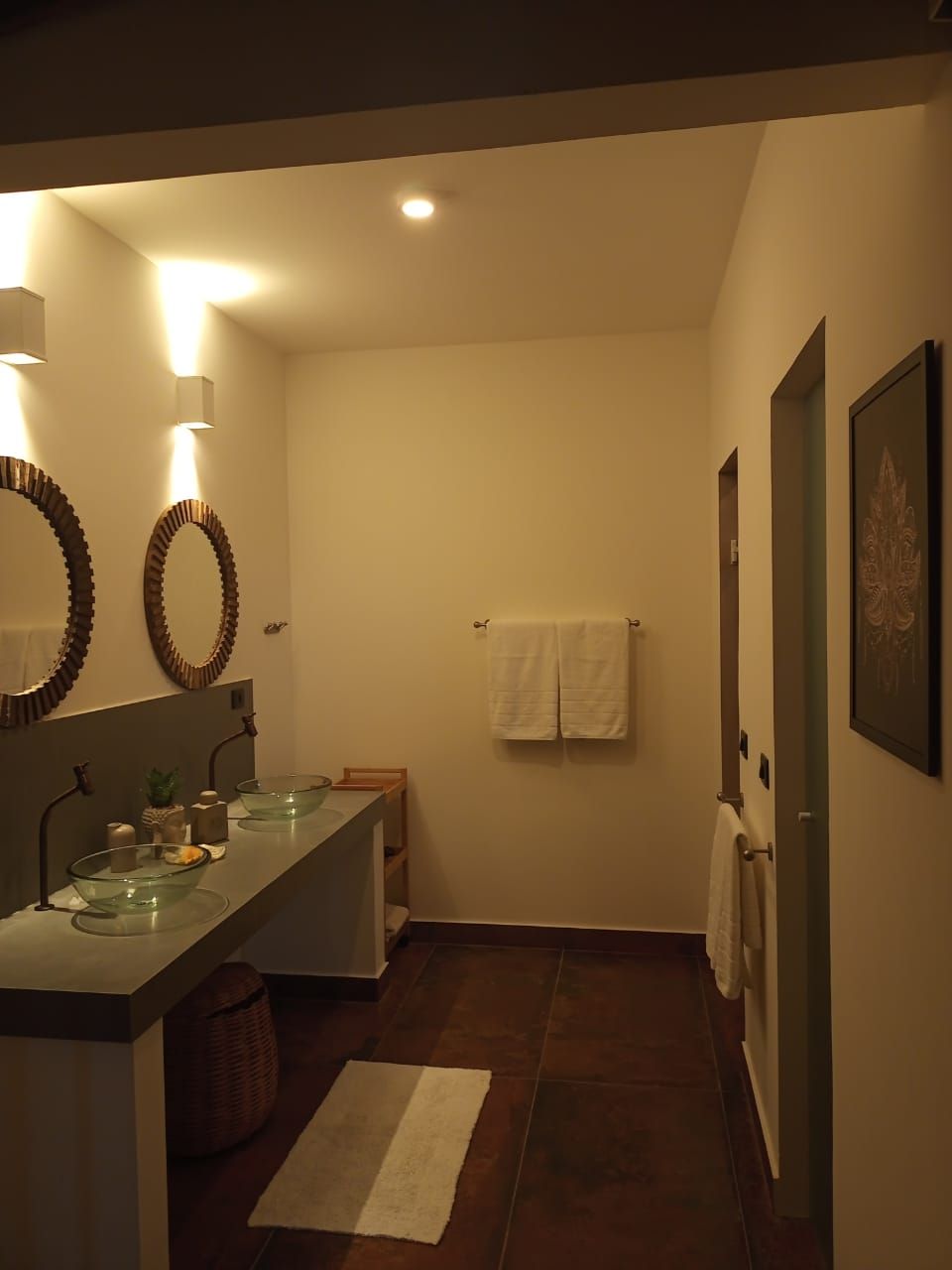 Un baño con un lavamanos de vidrio, dos espejos montados en la pared, un perchero con dos toallas, un cuadro enmarcado y un suelo de baldosas oscuras.