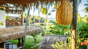 A thatched-roof outdoor area with a hanging woven basket, a wooden bench, and a view of a lush garden and a building in the background. - Hara Pousada e SPA (São Miguel do Gostoso)