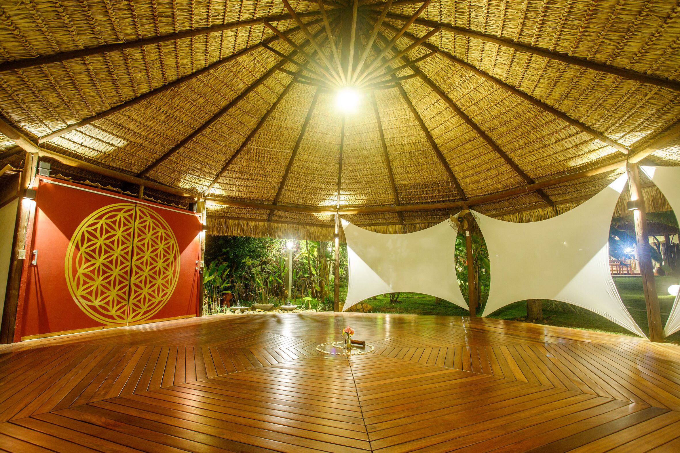 An indoor space with a wooden floor, a thatched roof, and a large circular design on the wall.