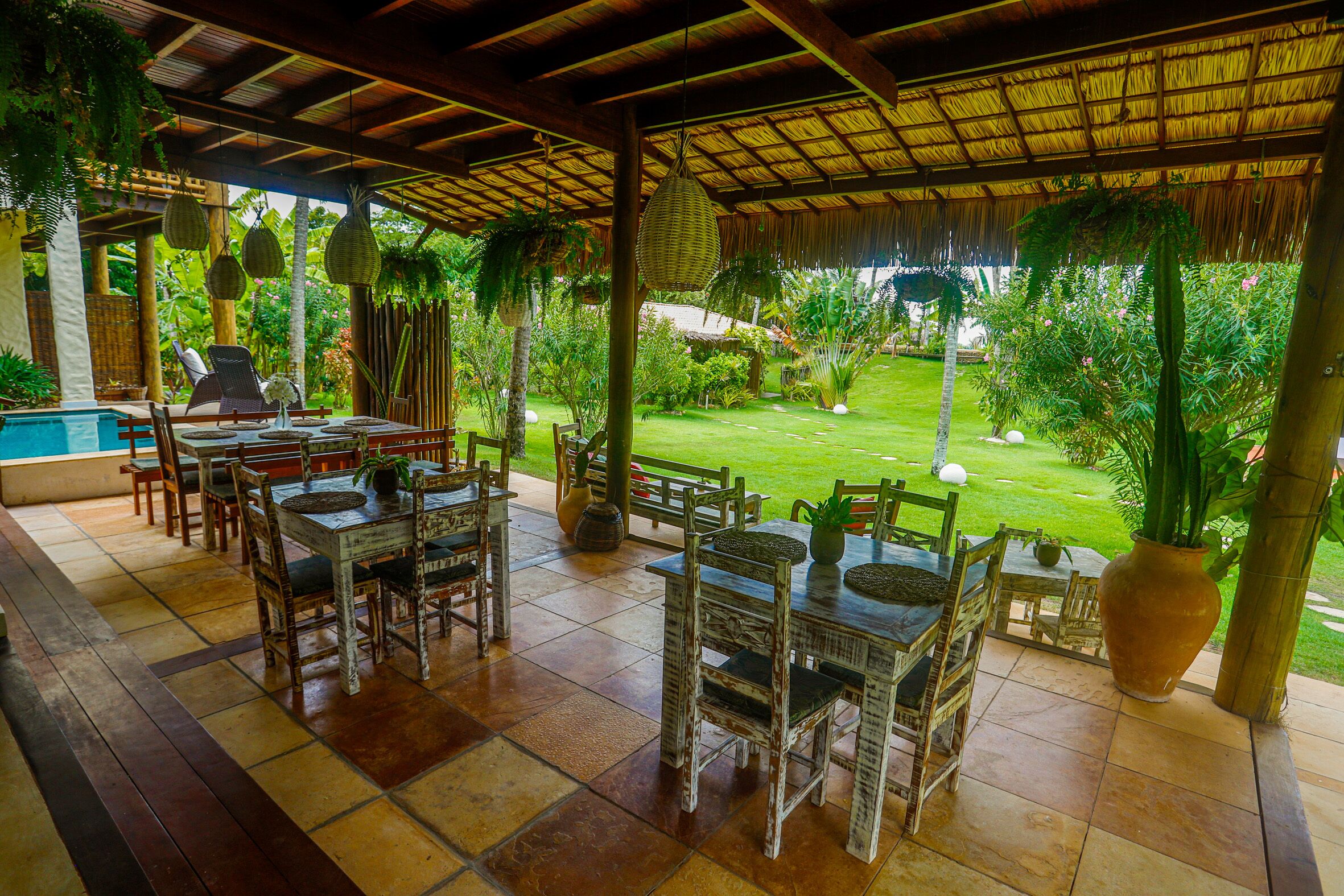 An outdoor dining area with wooden tables and chairs, surrounded by lush greenery and a thatched roof.