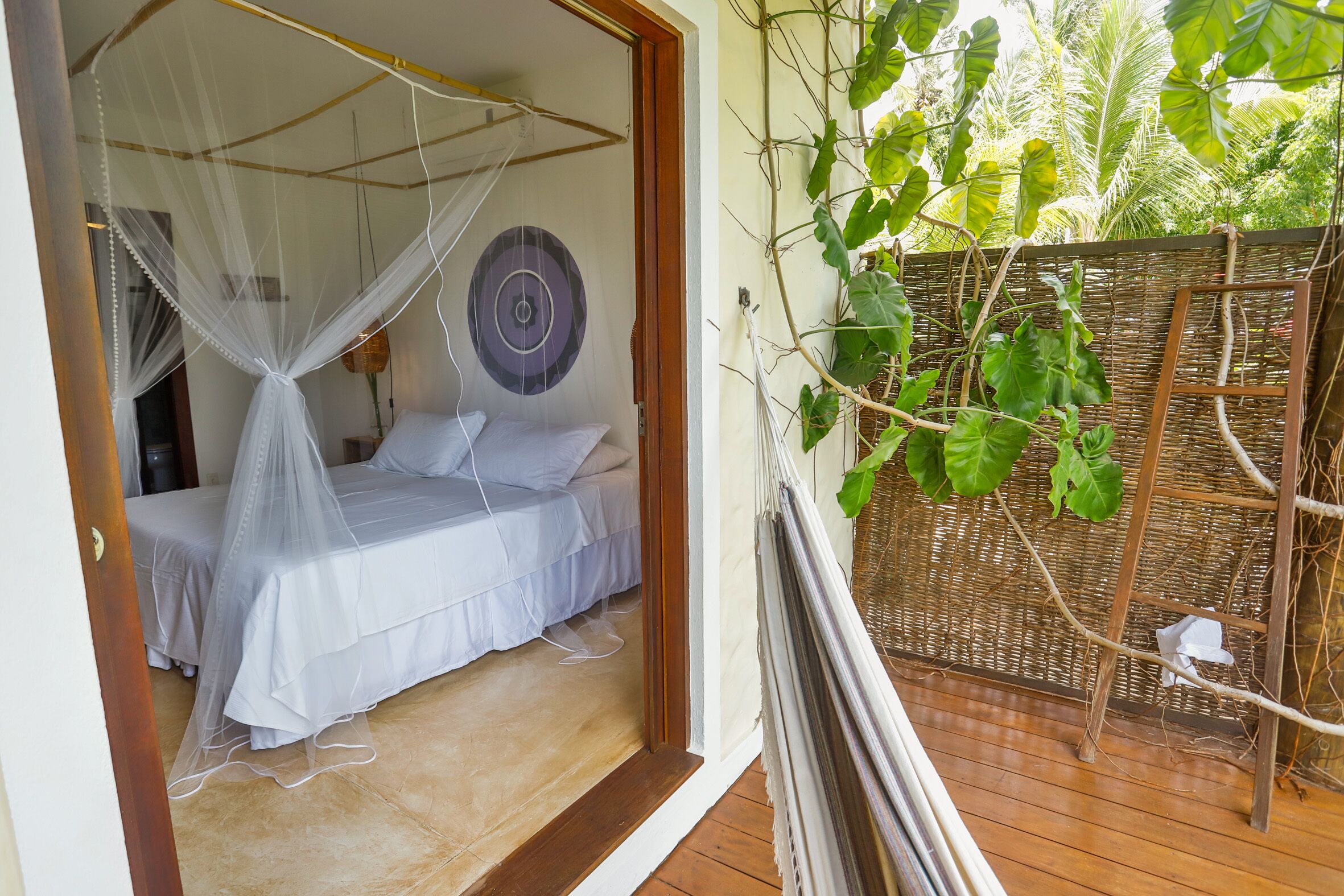 A bedroom with a bed under a mosquito net, a wooden floor, and a view of greenery outside.