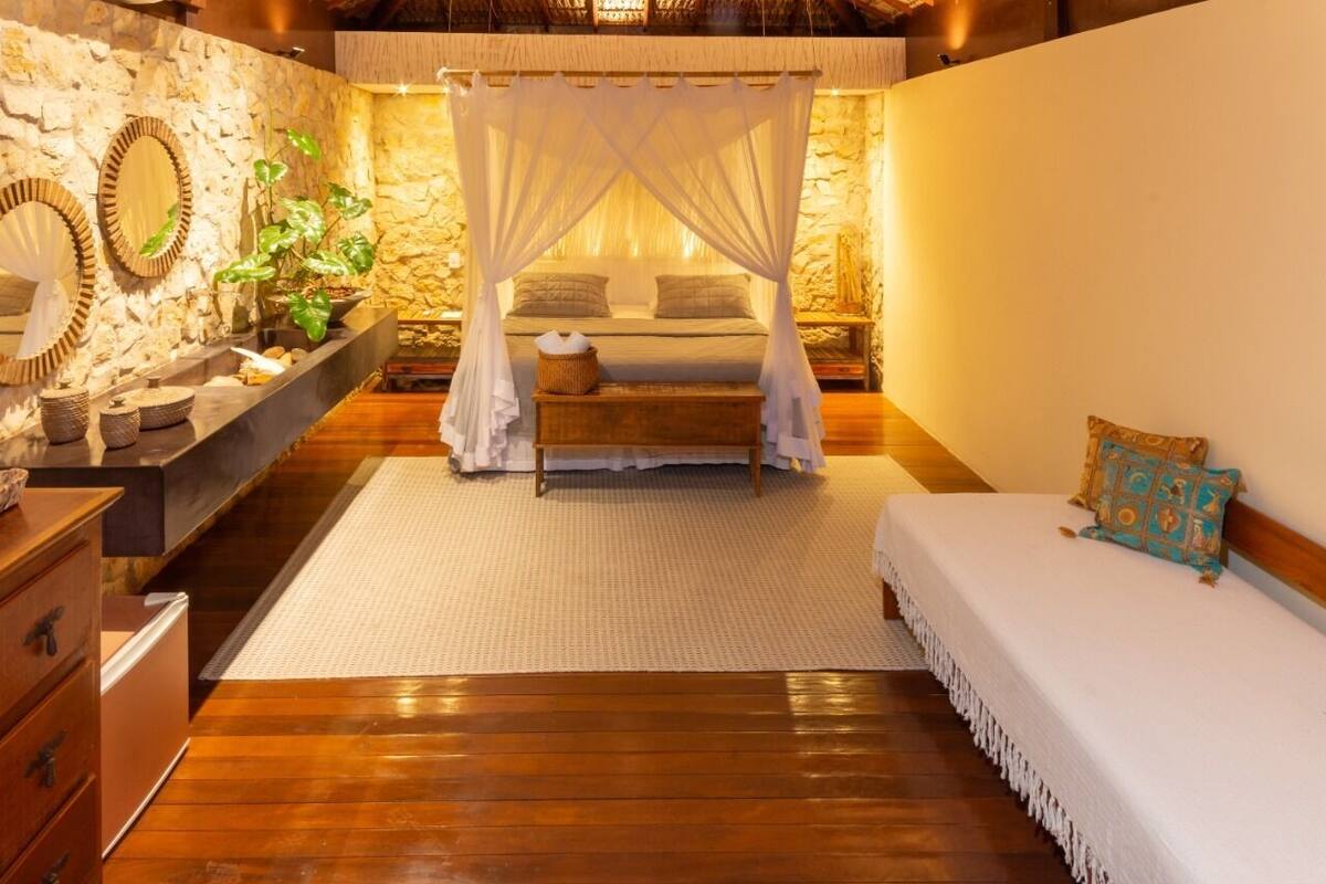 A bedroom with a canopy bed, wooden flooring, a stone wall, and a small bedside table with a plant.