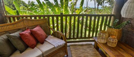 Eine Rattan-Couch mit Kissen, eine gewebte Matte und Körbe aus Rattan auf einer Holzterrasse mit Blick auf Palmen und das Meer.