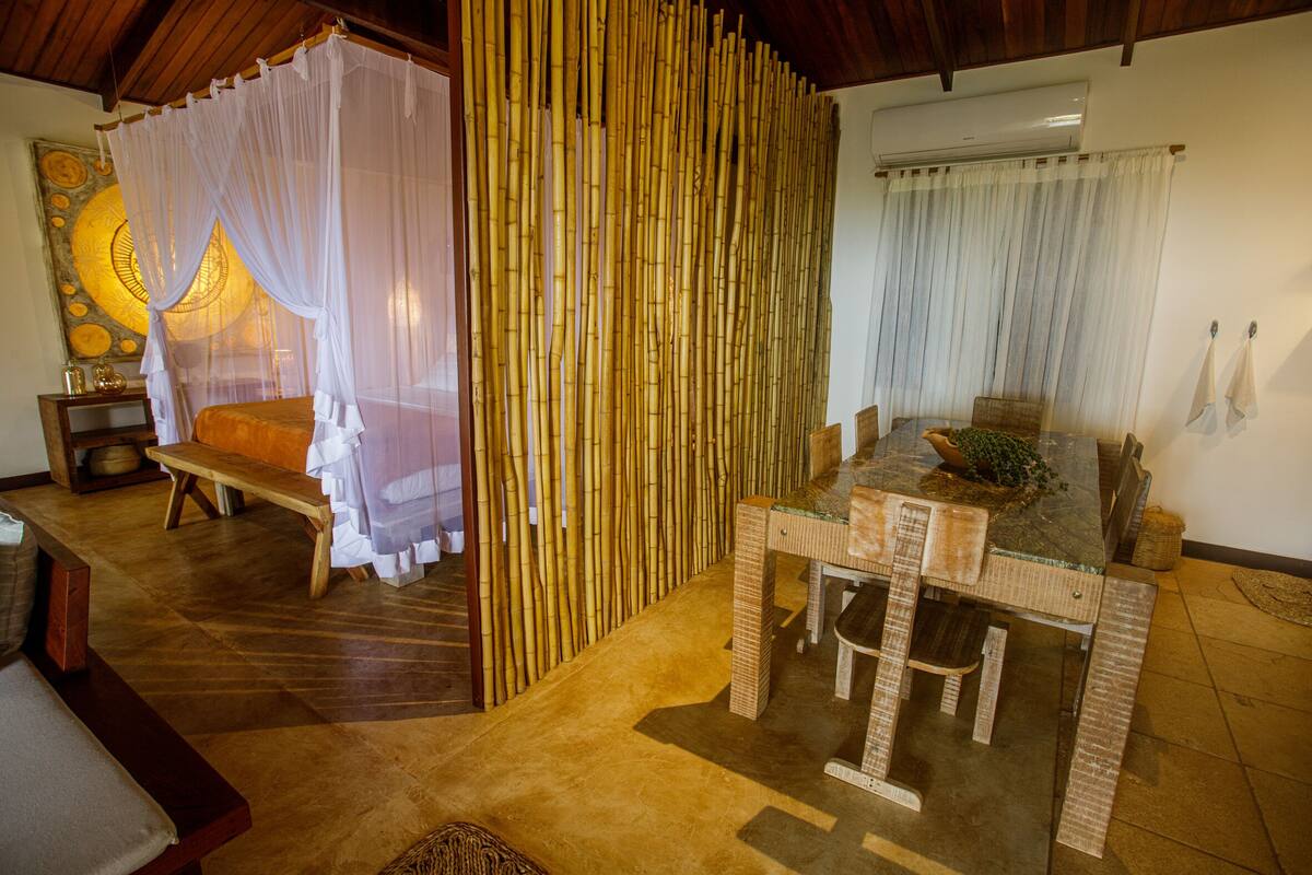 A room with a wooden floor, a glass-top table with chairs, a bench, a bed with a canopy, and a wall with a decorative circular pattern.