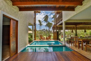Un área de piscina con dos sillones, una mesa de comedor con sillas y un espacio de estar cubierto.