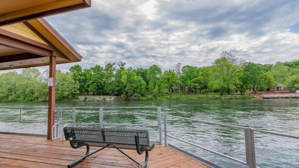 A wooden deck with a metal bench overlooking a river, surrounded by greenery and trees. - Bright and Gorgeous! Fish across the street - Lake Access in Branson! (Branson)
