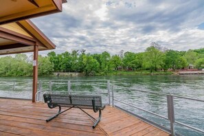 A wooden deck with a metal bench overlooking a river, surrounded by greenery and trees.