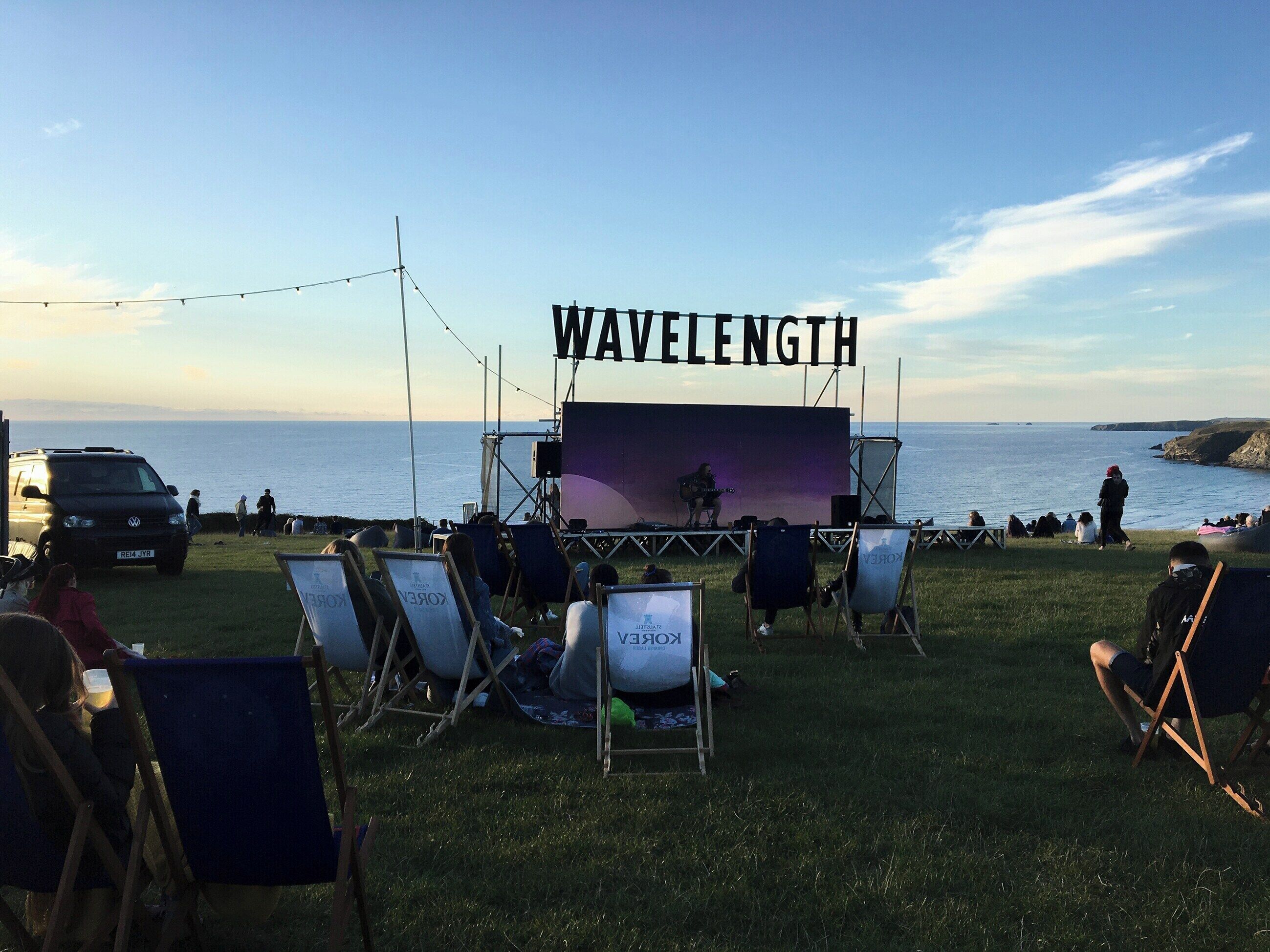 An outdoor stage with a backdrop reading 'WAVELENGTH' set against a coastal backdrop with grass seating and a van in the foreground.