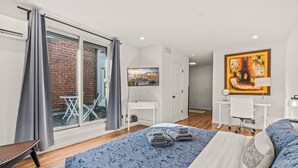 Ein modernes Schlafzimmer mit einem großen Bett, einem Schreibtisch und Blick auf ein Backsteingebäude durch eine Glasschiebetür.