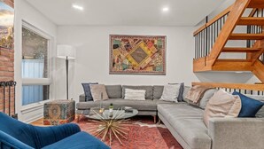 Ein modernes Wohnzimmer mit einem grauen Sofa, einem blauen Sessel und einem Couchtisch. Ein farbenfroher Teppich und ein großes abstraktes Gemälde sind an der Wand.