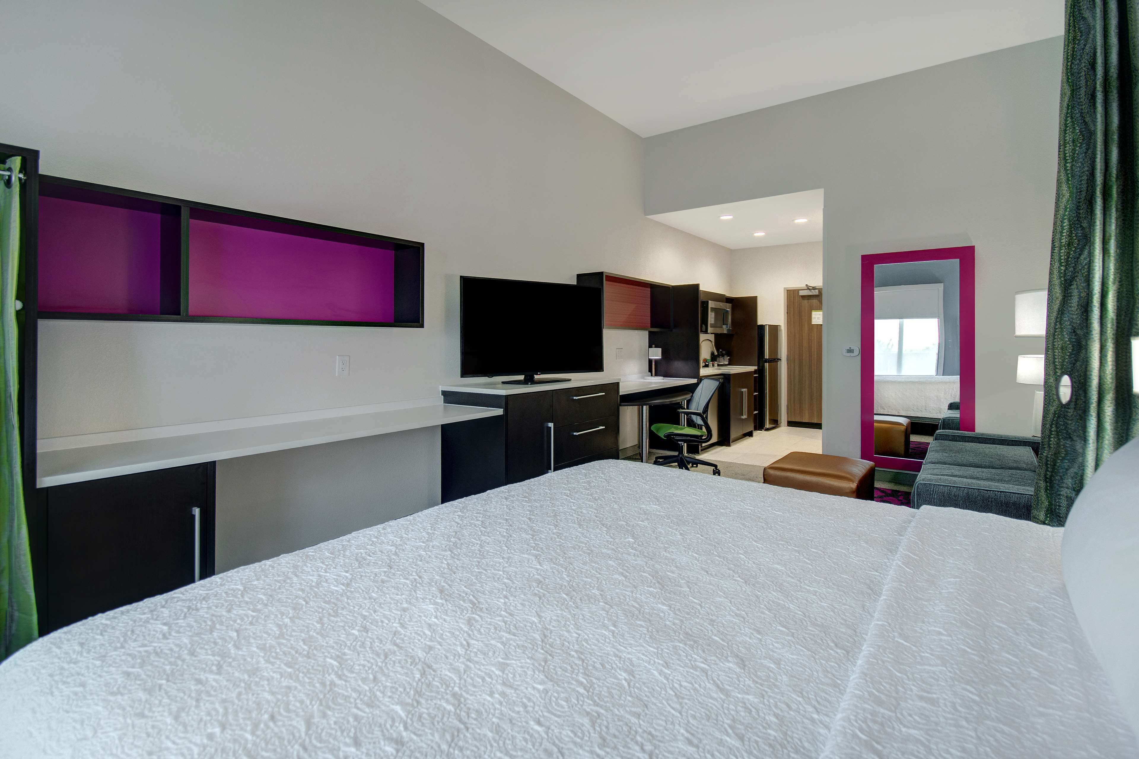 A modern hotel room with a large bed, a desk, a flat-screen TV, a kitchenette, and a bathroom with a mirror.