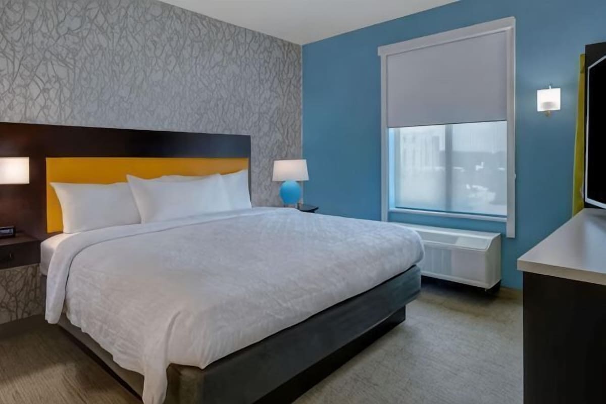 A hotel room with a large bed, a nightstand, a lamp, a window with blinds, and a wall with a textured pattern.