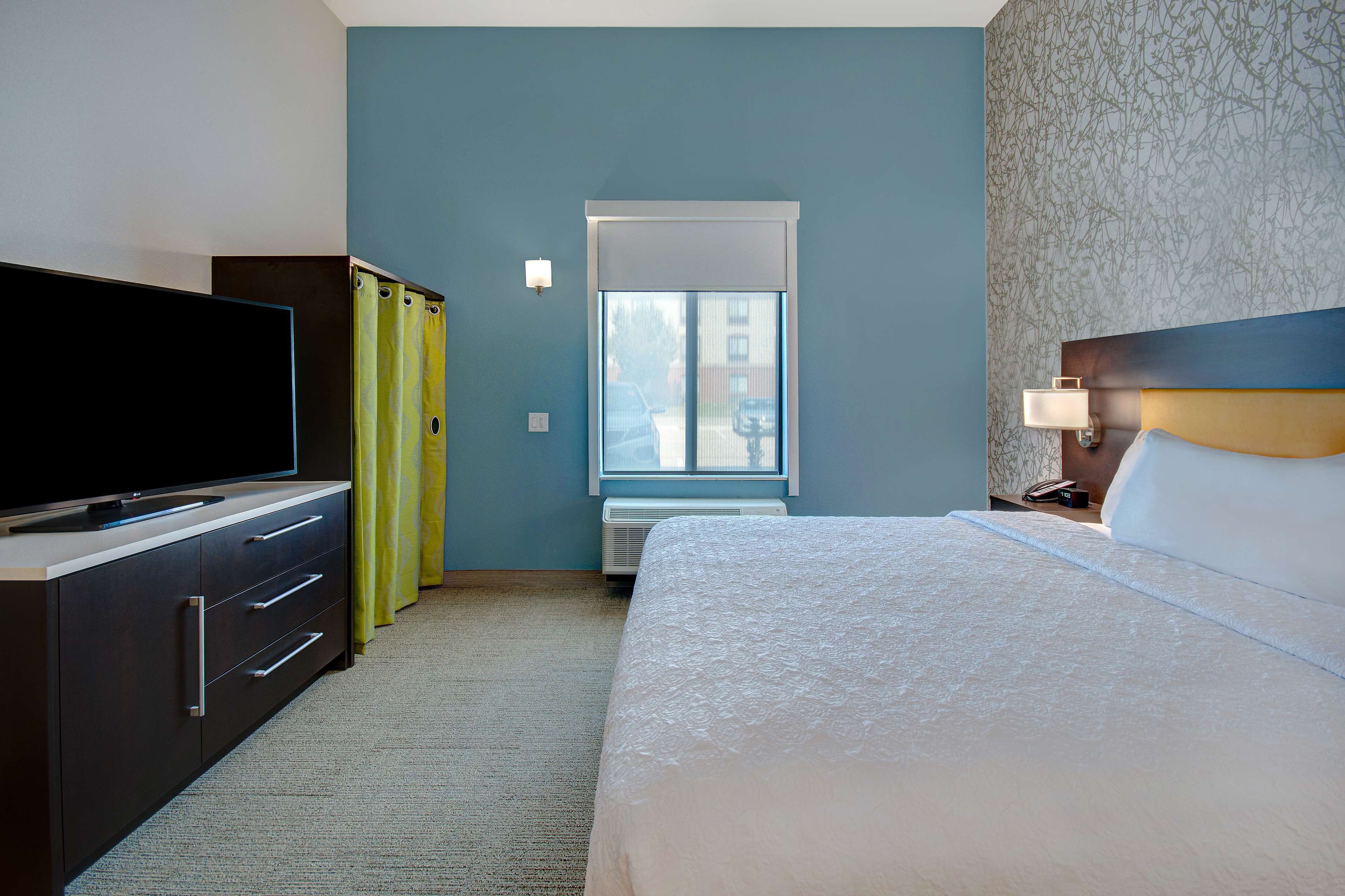 Una camera d'albergo con un letto, una TV su un comò, una finestra con tende e un comodino con una lampada.