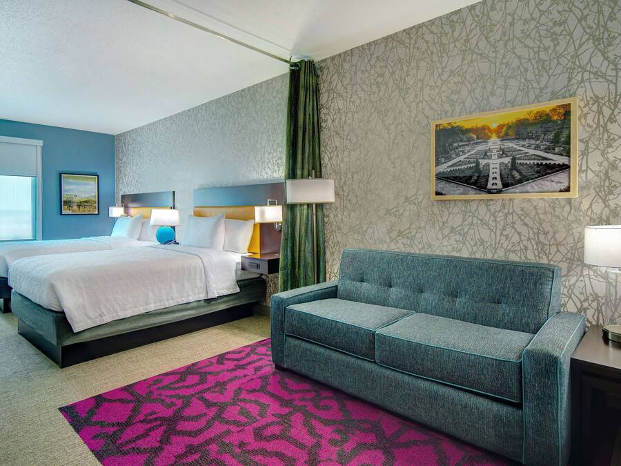 A hotel room with a bed, a sofa, a desk with a lamp, a picture on the wall, and a patterned rug.