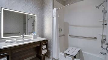 Un baño moderno con un espejo grande, un lavamanos, una ducha con cabezal de ducha de mano y un pequeño taburete.