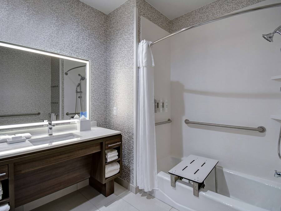Un baño moderno con un espejo grande, un lavamanos, una ducha con cabezal de ducha de mano y un pequeño taburete.