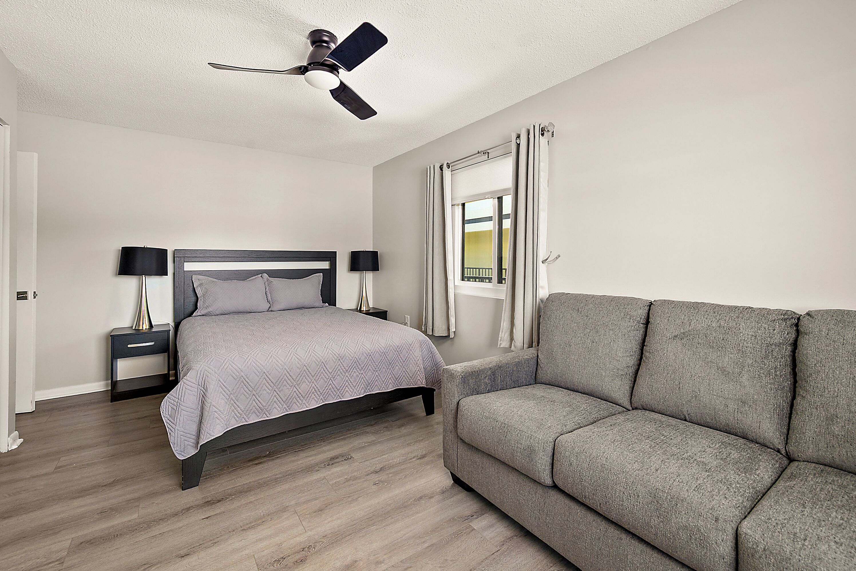 Quarto com cama, mesas de cabeceira, ventilador de teto, janela com cortinas e um sofá cinzento.